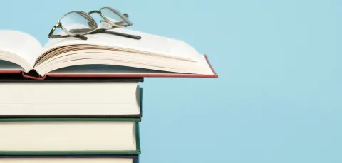 A stack of hardcover books with an open book on top and glasses resting on it against a blue background.
