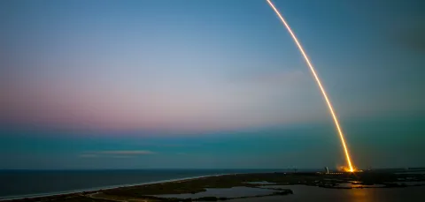 A rocket launch as seen from a distance at dawn or dusk. This is a long exposure that shows the launch as an arc of light from the ground into the sky