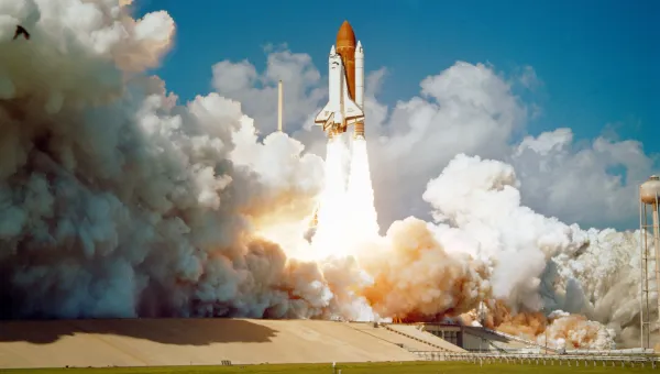 A space shuttle lifts off into the sky, with bright flames and thick smoke coming from its engines. The launch pad is visible below, and there are a few clouds in the blue sky.