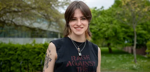 A student stands smiling outside the Physics building dressed casually in a sleeveless band T-shirt.