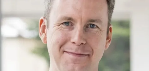 A man with short hair wearing a light grey sweater, standing in front of a window, with blurred greenery and wooden beams visible in the background.