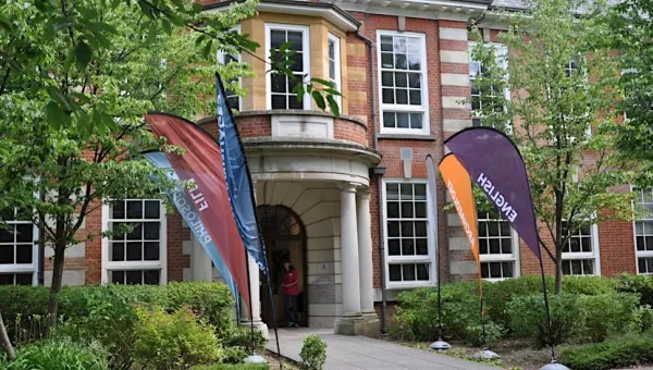 A brick building with large white-framed windows and a columned entrance is surrounded by greenery. Colorful flags labeled “English” and “Film” stand along the pathway leading to the doorway.