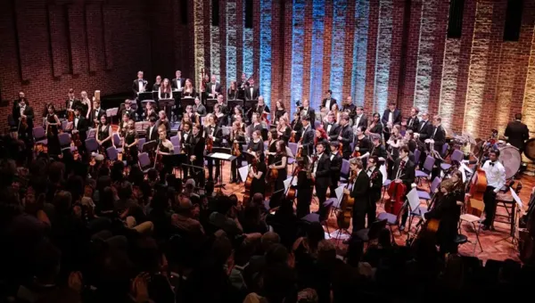 A full orchestra stands on stage in a concert hall with brick walls and vertical lighting accents, facing an audience seated in the foreground. Musicians hold various instruments including strings, woodwinds, brass, and percussion, with music stands arranged across the stage.
