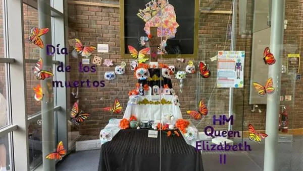 A Día de los Muertos altar is displayed behind glass, decorated with colorful paper butterflies, sugar skulls, flowers, and candles. Text on the glass reads “Día de los muertos” and “HM Queen Elizabeth II.”