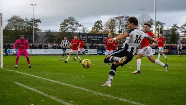 A football player in black‑and‑white kit stretches to strike a ball near the goal, while opponents in red attempt to block and the goalkeeper prepares to react.