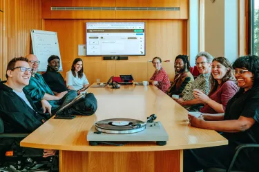  A group of nine people is seated around a wooden conference table, smiling and engaged in a meeting. Laptops are open, and a turntable with a vinyl record sits at the front of the table. A large screen behind them shows a web interface, and a whiteboard with notes is visible. The mood is friendly and collaborative in a modern, well-lit room.