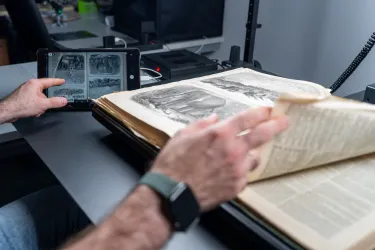 A close-up of a person's hands working with an open historical book on a specialized scanning table. One hand is turning a delicate page with illustrations, while the other holds a tablet displaying a digital scan of the same page. The workspace includes modern scanning equipment and cables in the background, blending traditional and digital technologies.