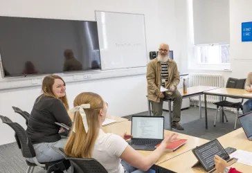 Alt text: A teacher sits on a desk holding papers while three students face them, one using a laptop. A whiteboard behind the teacher displays the phrase "POINT OF VIEW."