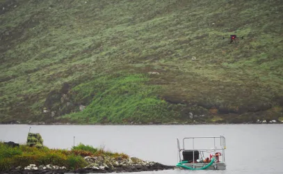 A drone hovers over a lake near a small floating platform with equipment, surrounded by lush green hills. The scene suggests a scientific or environmental study in a remote natural location.