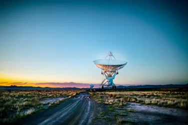 A towering satellite dish positioned in a wide, grassy field, silhouetted against a vivid orange sunset sky, with a winding dirt road leading toward it.