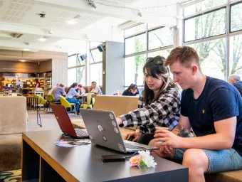 Two students sat working on laptops in a large, airy cafe.