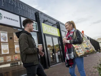 2 students with coffee standing outside the main entrance to the Piazza food court.