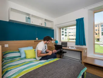 A student sits using a tablet on a large, brightly coloured bed in a modern bedroom. Modern buildings and a grassy courtyard can be seen through the windows.