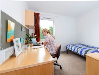 A spacious, bright student bedroom showing a single bed, desk and open window. A student sits at the desk, working on a laptop.