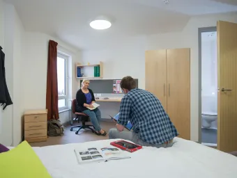 Two students sat talking in a large spacious bedroom. A double bed, desk and open door to a bathroom can be seen.