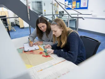 2 students studying a map at a table in a Waterfront Campus learning space. 