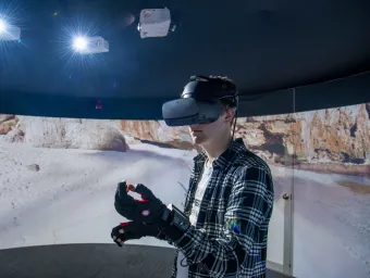 A student wearing a virtual reality headset and gloves, standing in front of a 360 degree video wall displaying what he can see.