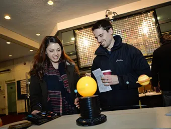 At the centre of the frame, there is a model of the sun, with other planets orbiting around it. Two young people are focused on the sun, and seem like they are enjoying themelves