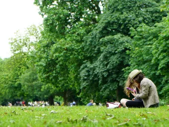 Two people sat down on the grass in a park looking at a book with trees and groups of people behind them. Stock image from pexels, by Chris J Mitchell.