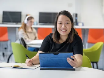 Jessie is sitting at a desk smiling at the camera. She has a tablet in 1 hand and a pen and note book in the other hand.