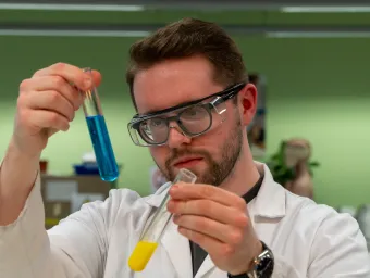 Nicholas in a lab coat and protective eyewear. He's holding two test tubes; one is filled with blue liquid, the other filled with yellow liquid.