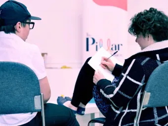 Two young adults sitting with their backs to the camera. One has their feet up on a table and is writing on a note pad resting on their knee. The other has a baseball cap on and is watching the person writing. there is a Pillar logo on the wall.