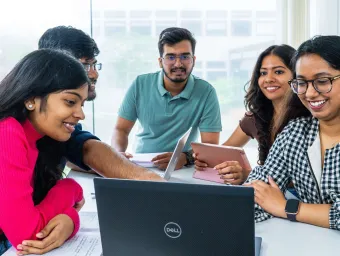 A group of university students work together at a table, smiling and using laptops and tablets.