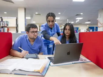 Medicine students wearing blue scrubs, study together in a library, with a laptop, tablet and reference book