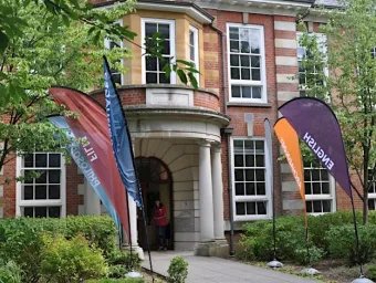 A brick building with large white-framed windows and a columned entrance is surrounded by greenery. Colorful flags labeled “English” and “Film” stand along the pathway leading to the doorway.