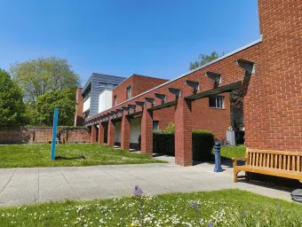 A courtyard with red brick buildings features a covered walkway supported by square pillars and a wooden bench on the right. The area is surrounded by green grass, small flowers, and trees under a clear blue sky.