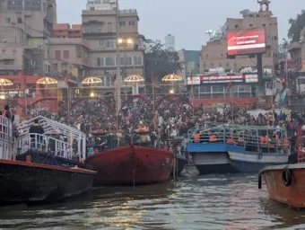 Evening view of Dashashwamedh Ghat in Varanasi, India, with large crowds gathered for the Ganga Aarti ceremony. Several colorful boats are docked along the banks of the Ganges River, facing the illuminated steps leading up to temples and buildings.