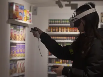 A person wearing a virtual reality headset and holding controllers interacts with a simulated environment that resembles a grocery store aisle. Shelves in the background display various colorful packaged food items.