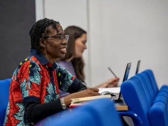 Ogene Adoh sat in a lecture classroom. She has a laptop and notebook in front of her and is looking towards the front of the classroom. 