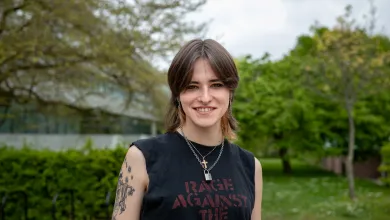 A student stands smiling outside the Physics building dressed casually in a sleeveless band T-shirt.