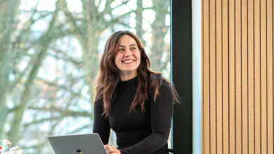 A student sitting on a window ledge seat smiling at the camera. She has a laptop on her knee and there are trees behind her.