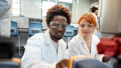 Two students wearing white lab coats and clear safety goggles are working together in a modern laboratory. The students are leaning over bench-top equipment adjusting a dial.