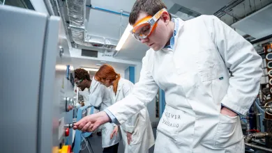 Student in a chemical engineering lab wearing a white lab coat and safety goggles adjusts controls on industrial equipment, while two other students work at a panel in the background.