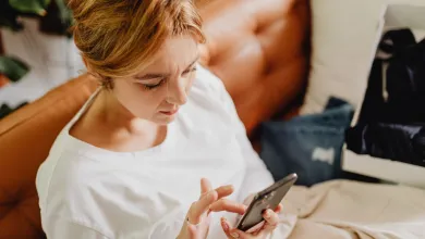 A person sits on a brown leather sofa indoors, holding and looking at a smartphone while wearing a loose white top. Soft natural light and houseplants in the background create a relaxed home setting.