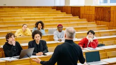 A lecturer stands at the front of a tiered lecture theatre addressing a small group of students seated at wooden desks with laptops and notebooks. Rows of empty benches extend behind them, and the room features wood‑panelled walls and bright natural light.