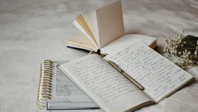An open notebook filled with handwritten notes lies on a soft surface with a pen resting across the pages, next to a spiral‑bound planner. An open blank book and a small bunch of dried flowers sit in the background under soft, diffused lighting.