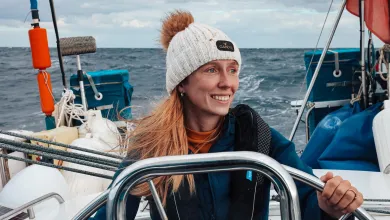 A person stands at the wheel of a small sailing boat on open water. The person wears a knitted hat with a pom-pom and a padded jacket.