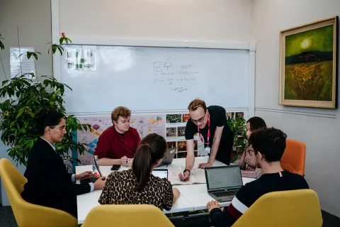A group of people is collaborating around a table in a modern office space, with laptops, papers, and a whiteboard displaying diagrams and notes. One person is leaning over, writing in a book, while others focus on their discussions and screens.