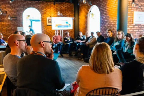 A group of people is seated in a circle. The participants are engaged in a discussion. A screen is visible in the background, displaying content