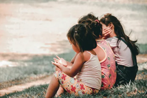 Three children sitting on grass, facing away, with colorful clothing.
