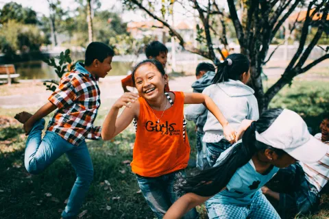 Children energetically playing outdoors, with a focus on a laughing girl in an orange shirt.