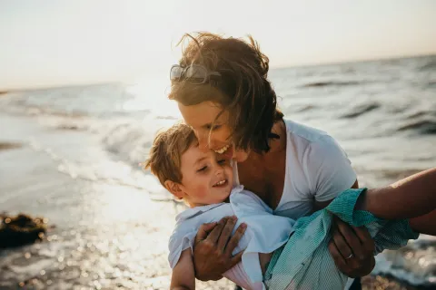 Woman holding a child on a sunny beach, both smiling.