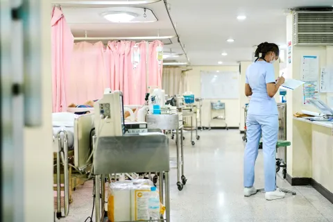A hospital ward showing a trolley of equipment at the end of each bed. There is 1 member of staff facing away from the camera completing a chart.