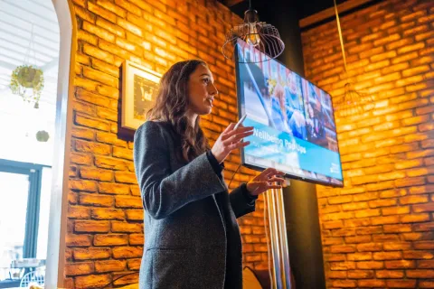 A woman is photographed from below, shown speaking in front of a digital screen.