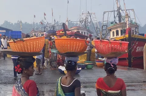 Three women at a ship harbour carrying baskets of fish on their heads.