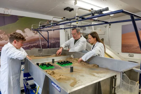 Three people in lab coats work at a sand-filled table with various objects and overhead nozzles, set against a backdrop of landscape images.